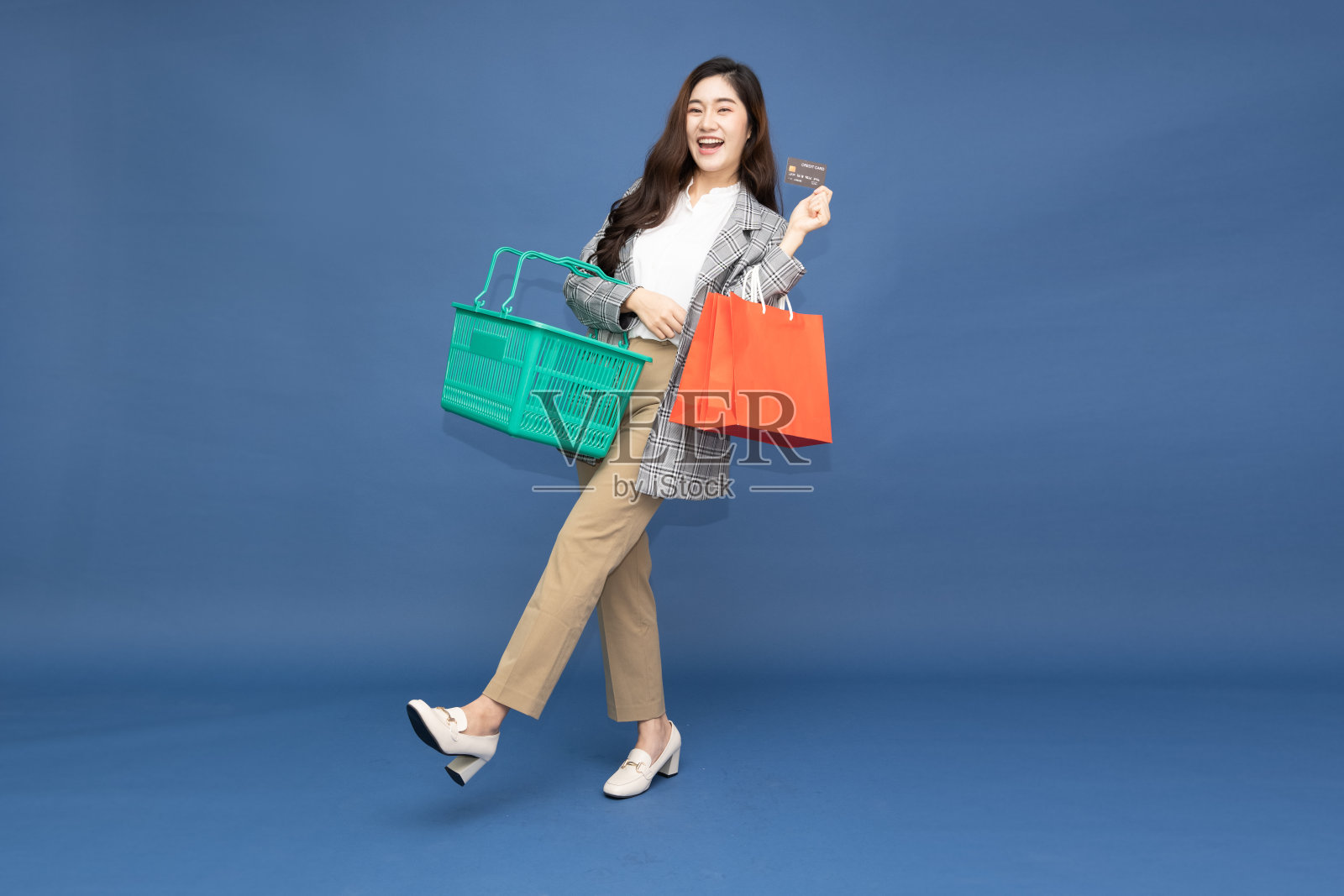 快乐漂亮的亚洲女商人在西装持有红色的购物袋，绿色的购物篮和信用卡孤立在蓝色的背景，全长肖像和购物概念照片摄影图片