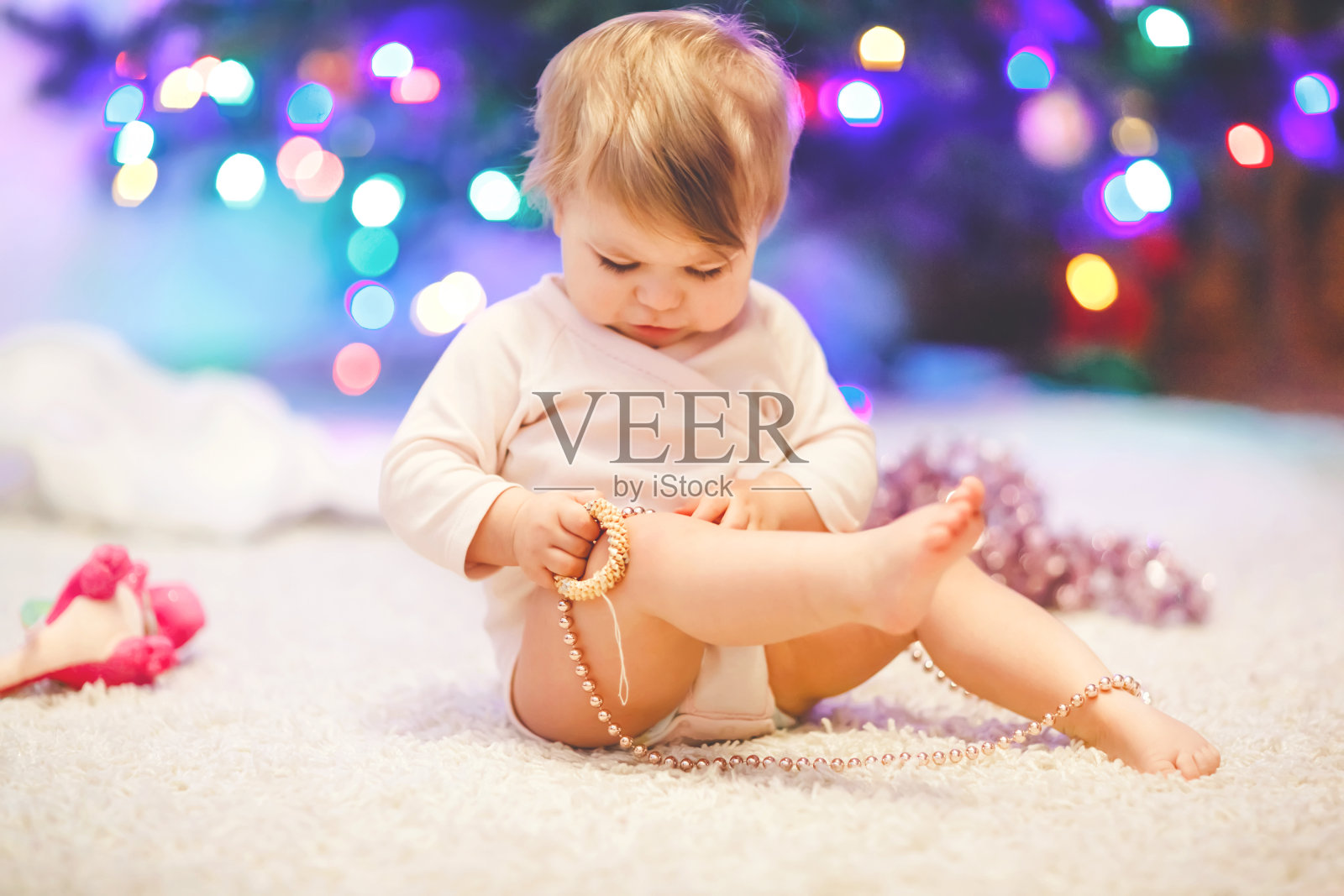 可爱的小女孩拿着粉红色的圣诞树珠子在可爱的手。小孩子和家人一起装饰圣诞树。第一次庆祝。蹒跚学步的孩子和彩色灯光的背景。照片摄影图片