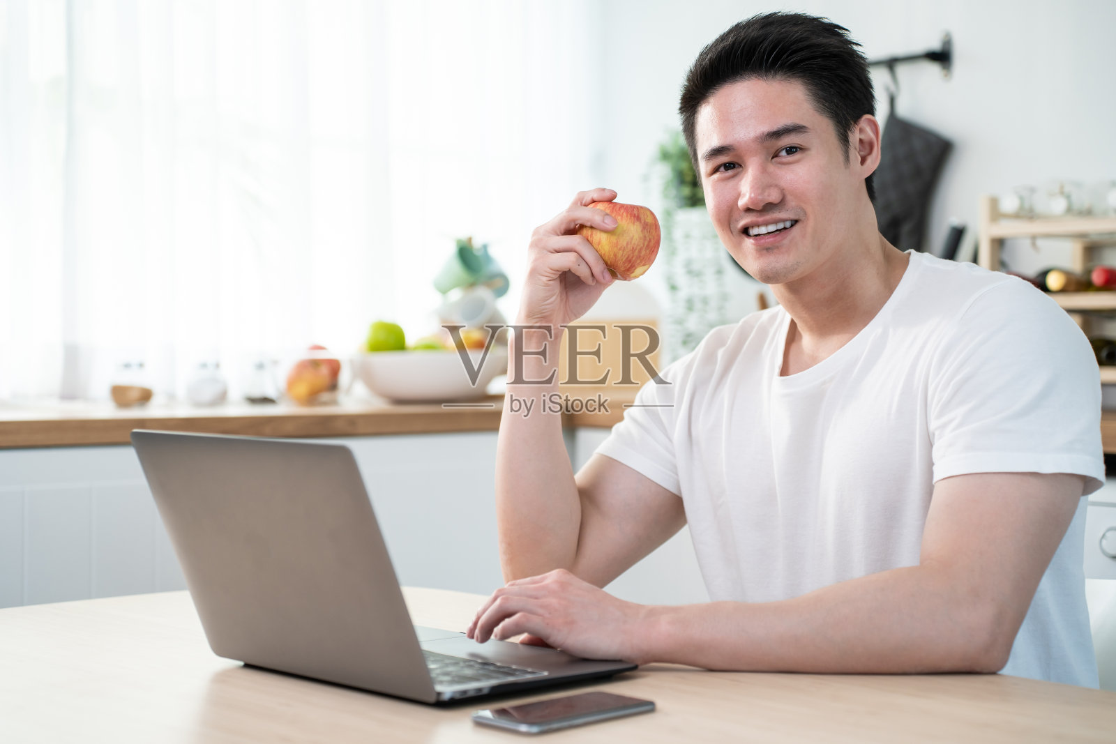 亚洲商人在家工作时吃健康沙拉的肖像。有魅力的帅哥用笔记本电脑上网学习，在厨房吃干净的蔬菜保健，看着相机。照片摄影图片